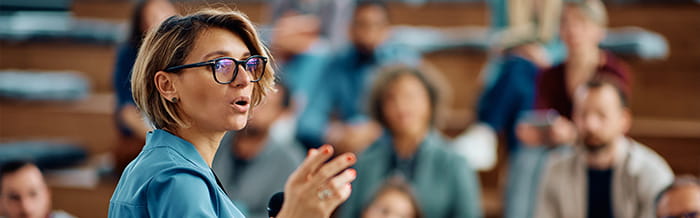 A female speaker with glasses and short blonde hair speaks emphatically during a conference, with an audience of blurred people sitting on bleachers behind her