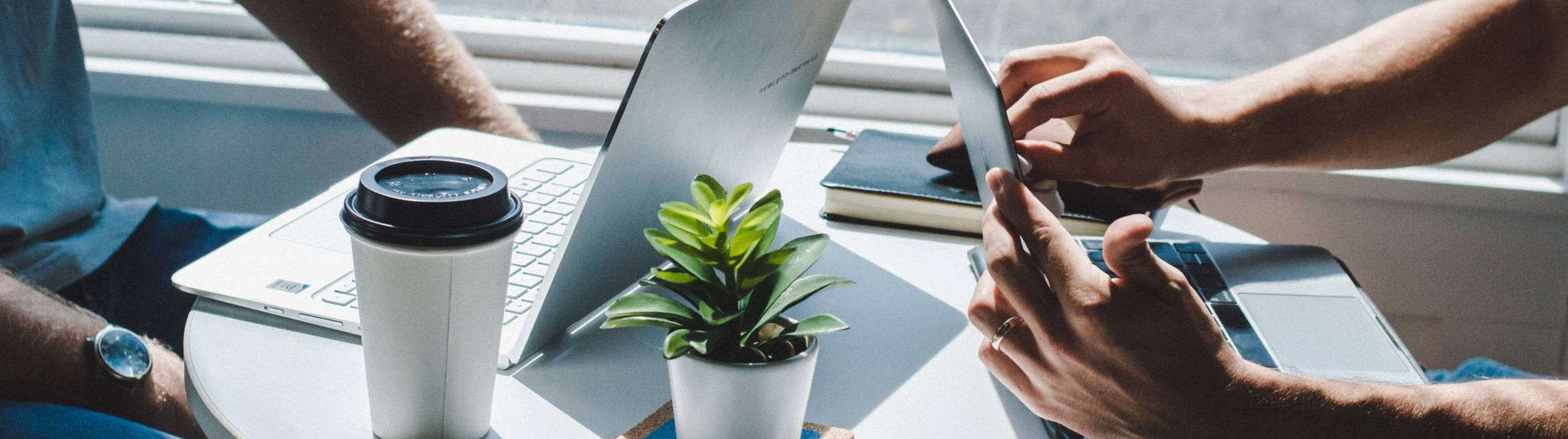 Two people sitting at a small circular table working on their laptops in a bright setting; a small potted plant, a notebook, and a takeaway coffee cup are on the table