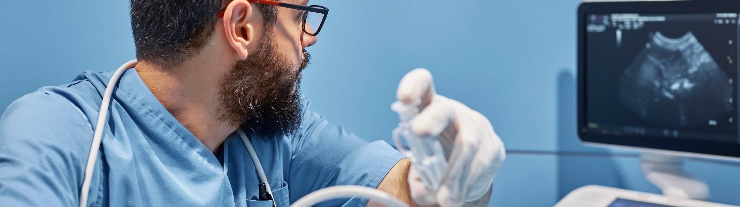 A veterinarian with a beard and glasses, seen in profile, carefully observes an ultrasound monitor while performing a diagnostic exam with gloved hands in a clinic with blue walls